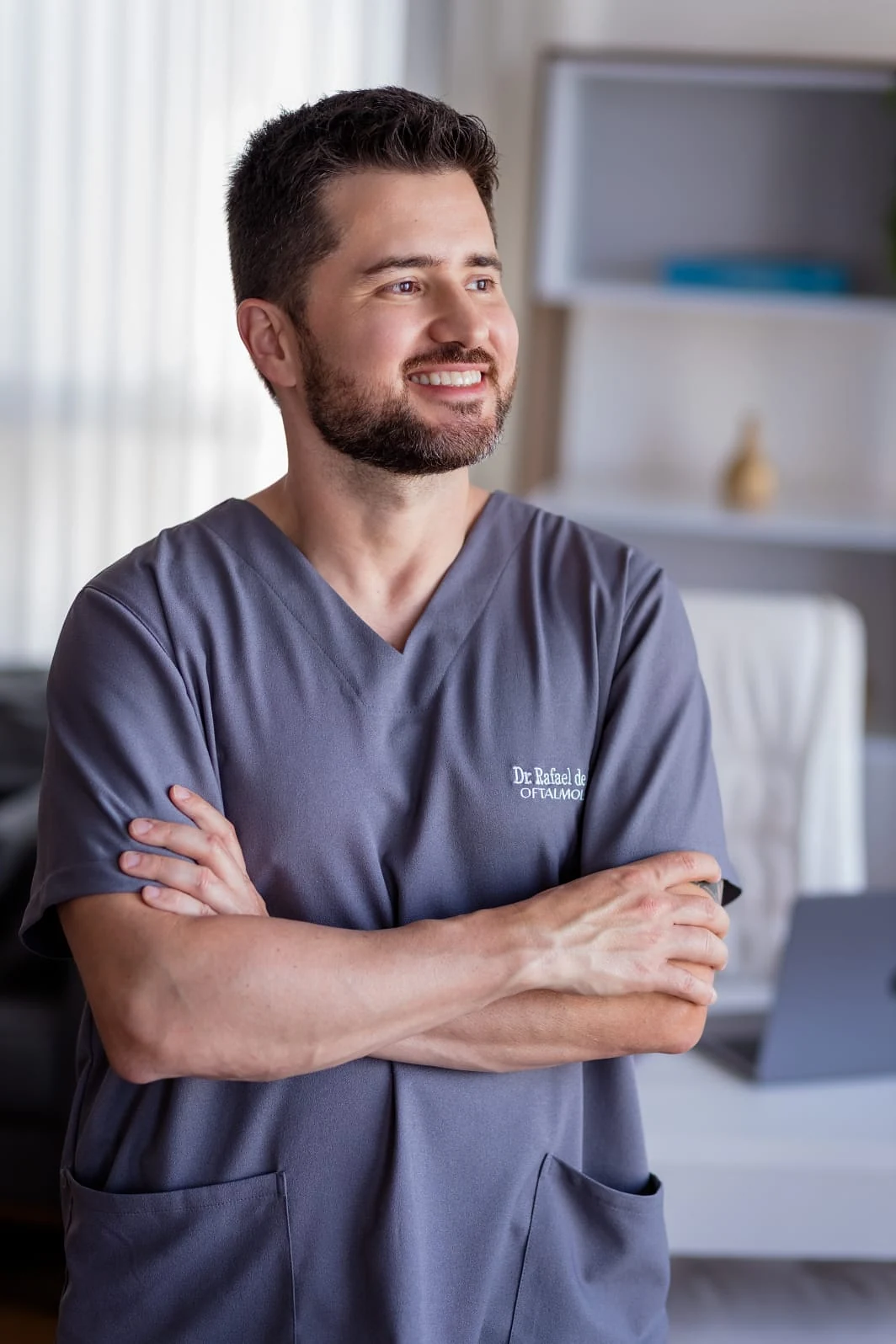 Dr. Rafael de Matos de pijama cirúrgico, de braços cruzados, sorrindo no consultório