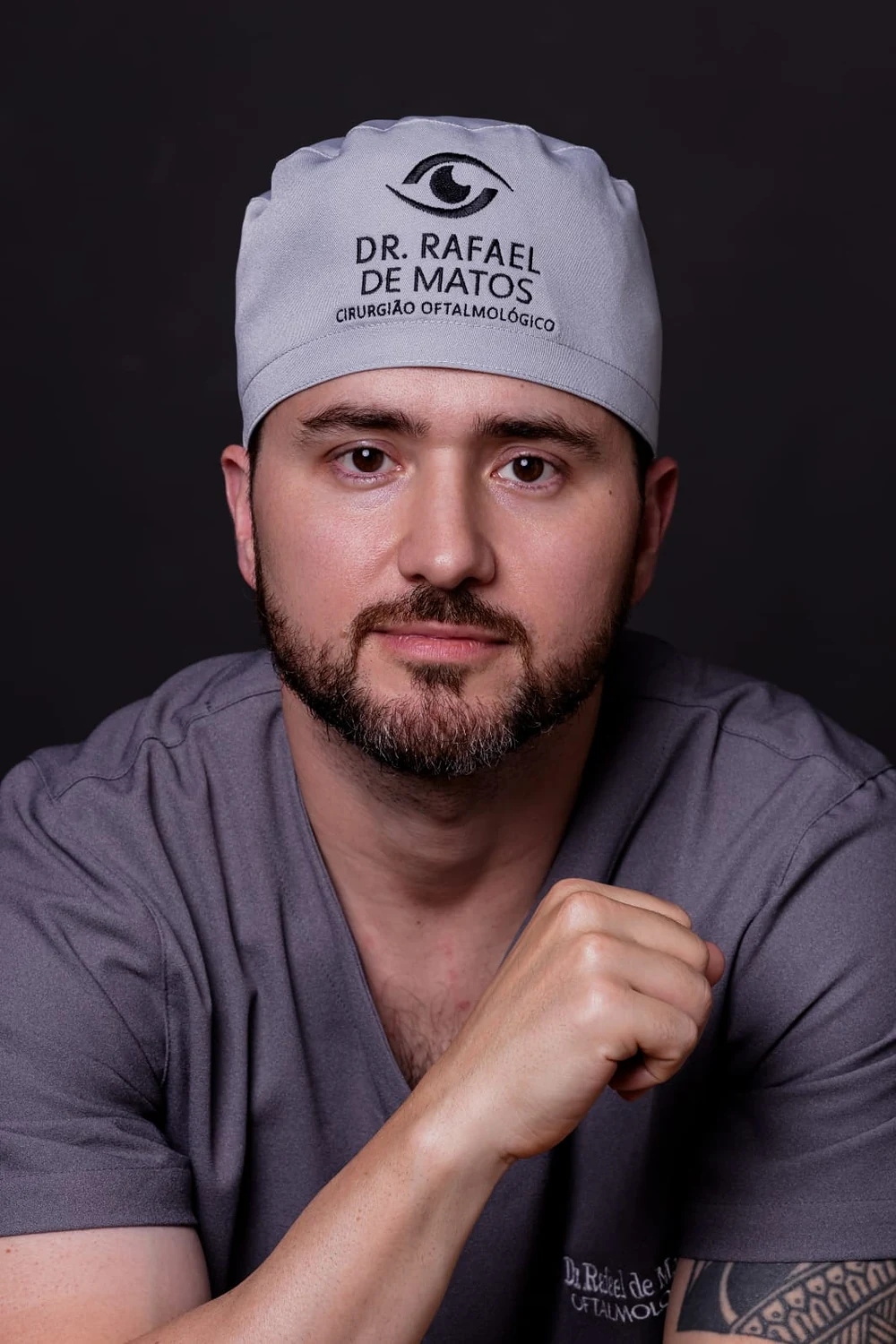 Dr. Rafael de Matos de pijama cirúrgico e touca com o seu nome, em fundo escuro